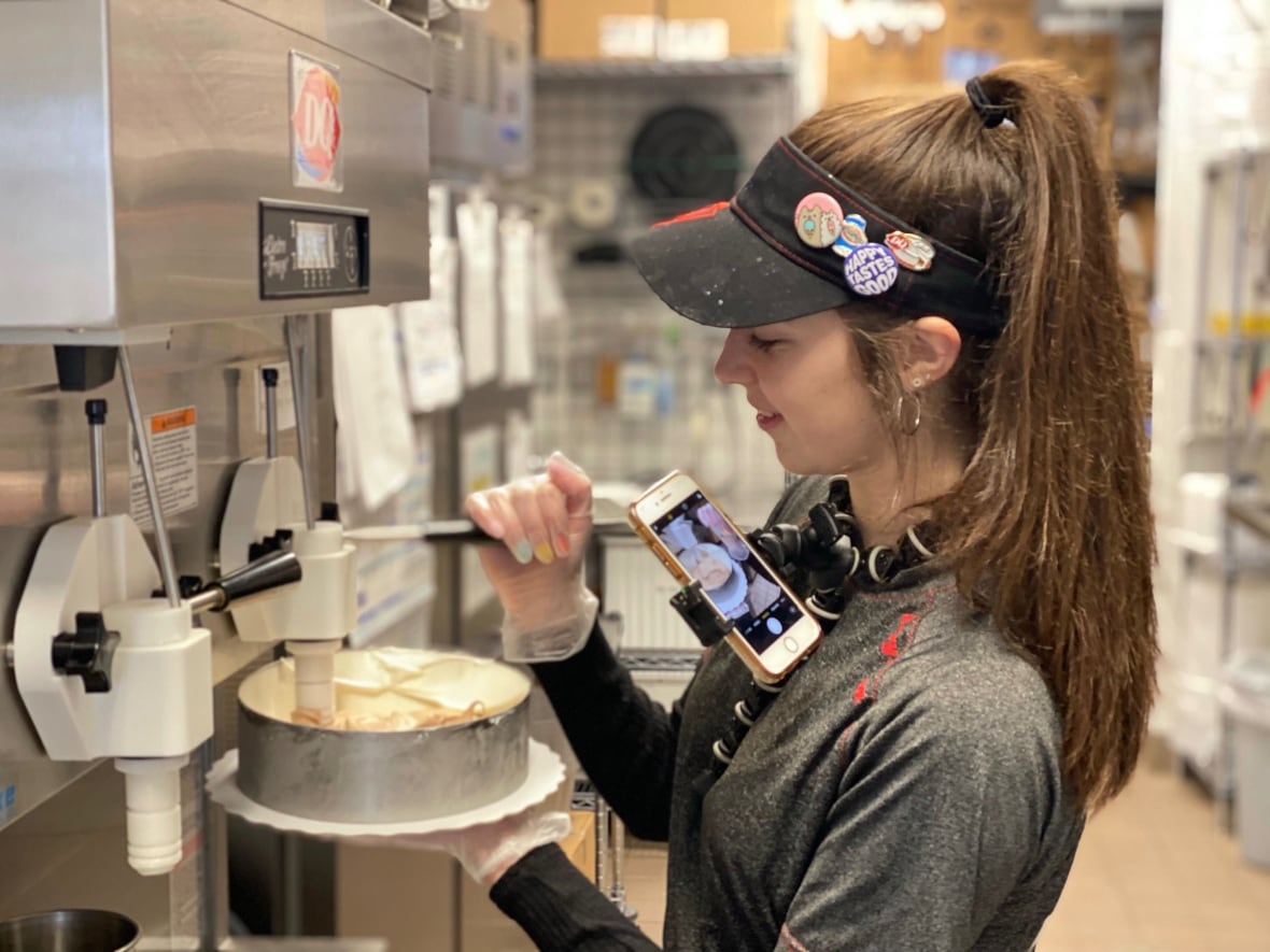 A woman wearing a hat puts ice cream in a cake mold while filming on her phone with a tripod.
