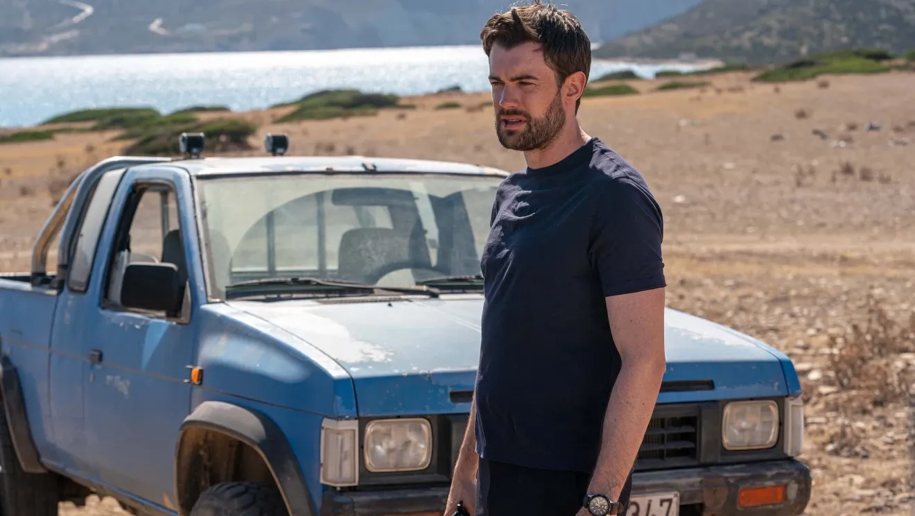 Jack Whitehall in a still from Malice. He's in front of a battered blue pickup near a lake