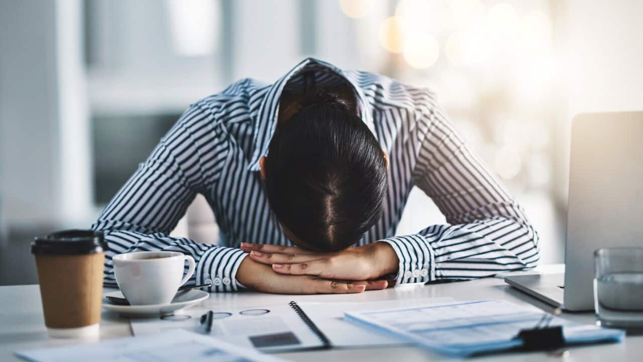 A woman with her head on her hands, pressed against a desk in an office. There is a laptop, files and coffee on the desk.