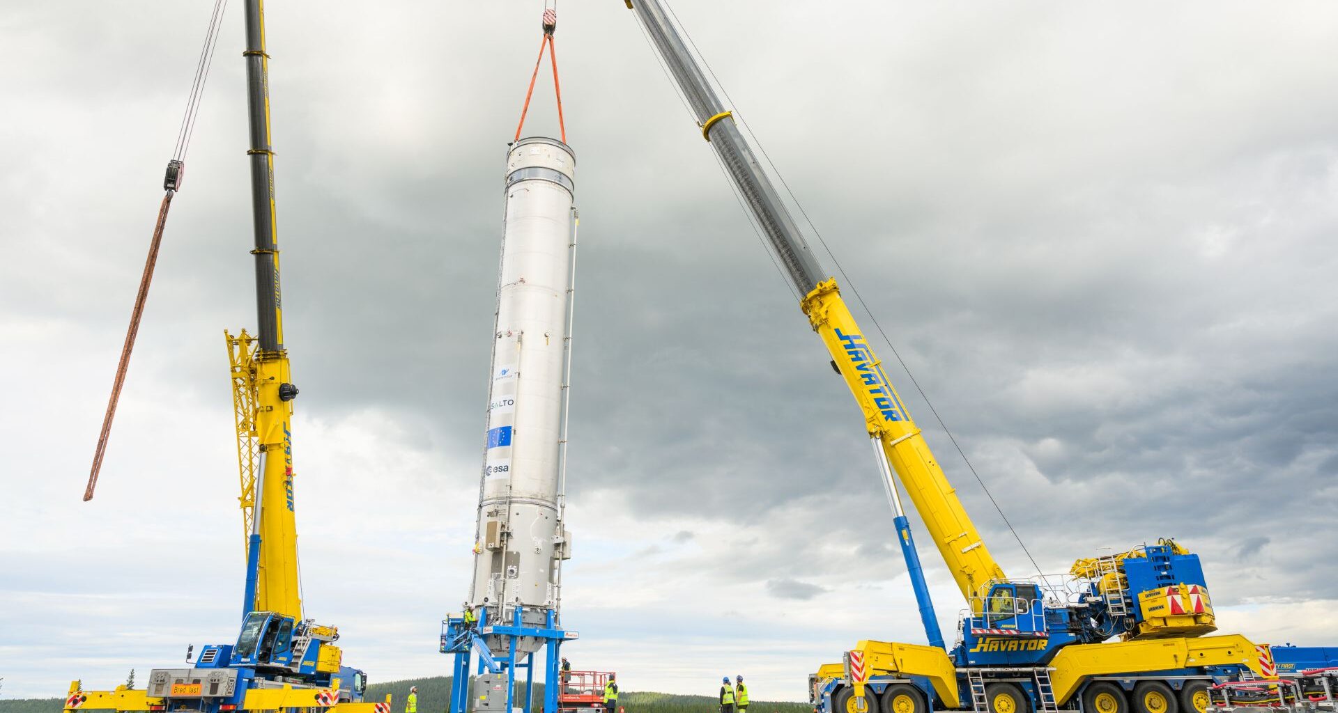 A white rocket is hoisted by yellow cranes