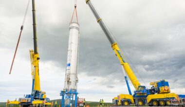 A white rocket is hoisted by yellow cranes
