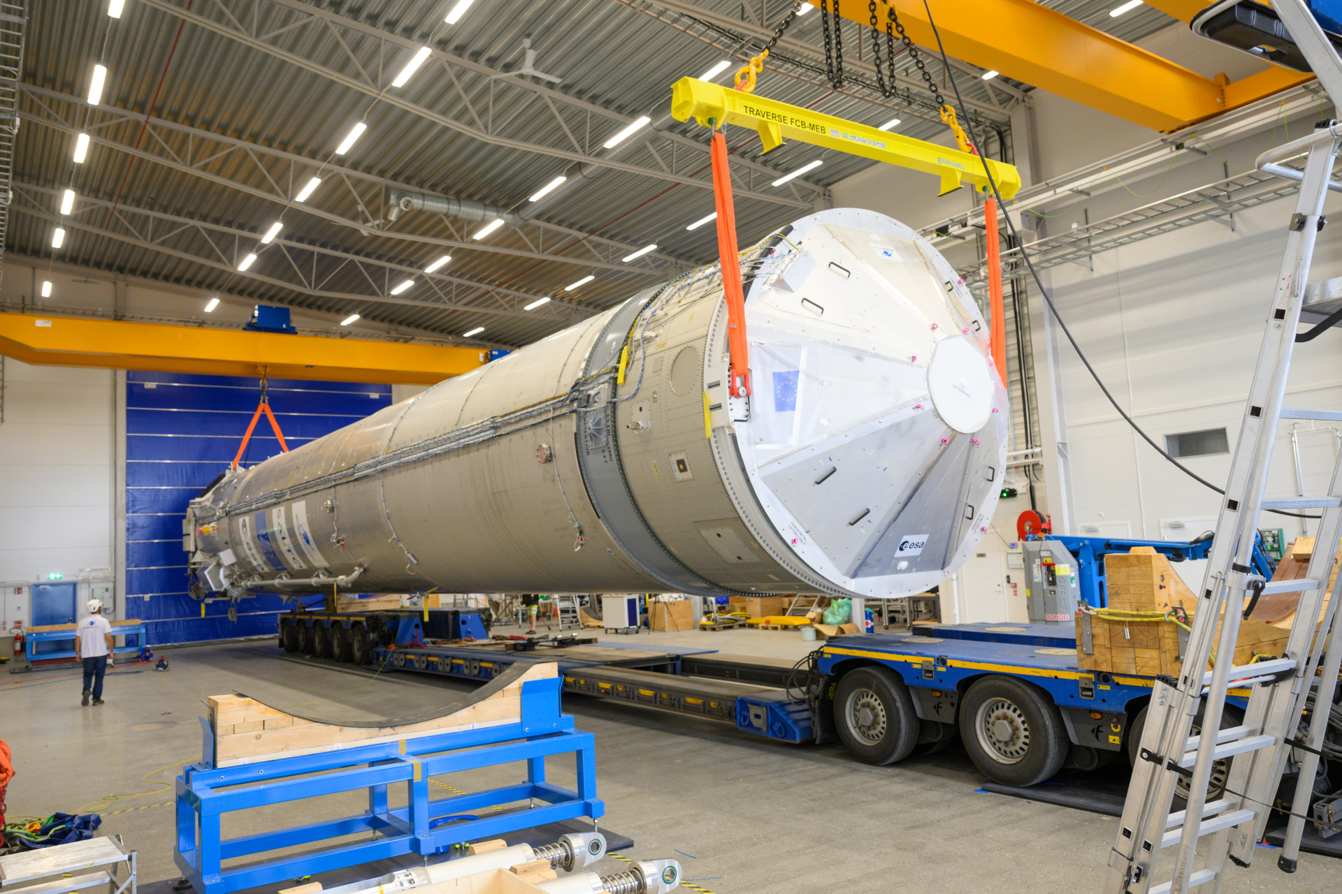 The white cylindrical body of a rocket is hoisted horizontally onto the flat bed of a truck.