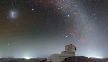 The domed roof of the Vera Rubin Observatory sits on a high ridge with a red and purple starry night sky above it with a glowing arch of the Milky Way seen in the heavens