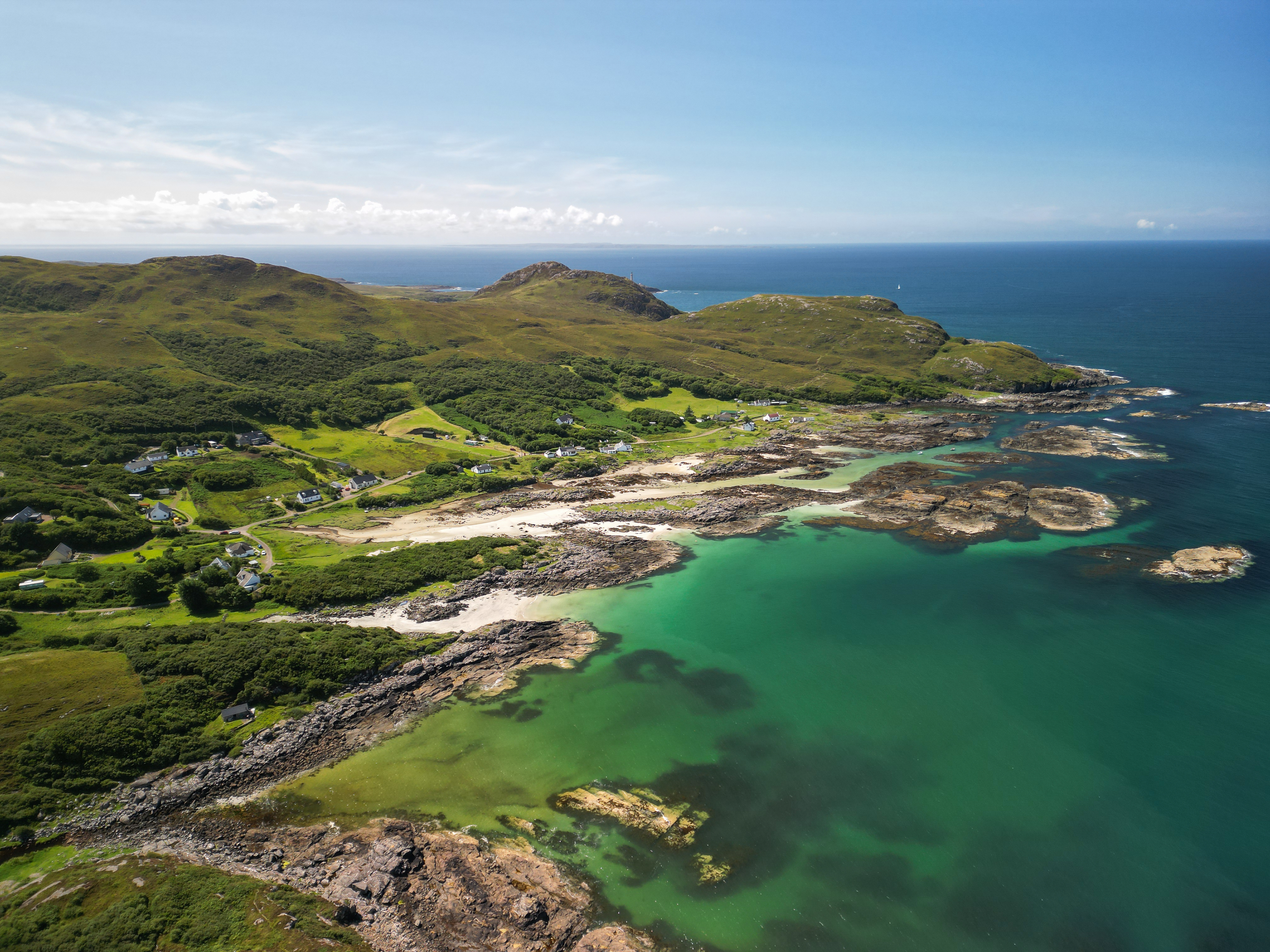 Aerial view of Sanna Bay in the Scottish Highlands, showing a sandy beach, green hills, small white houses, and clear turquoise water.