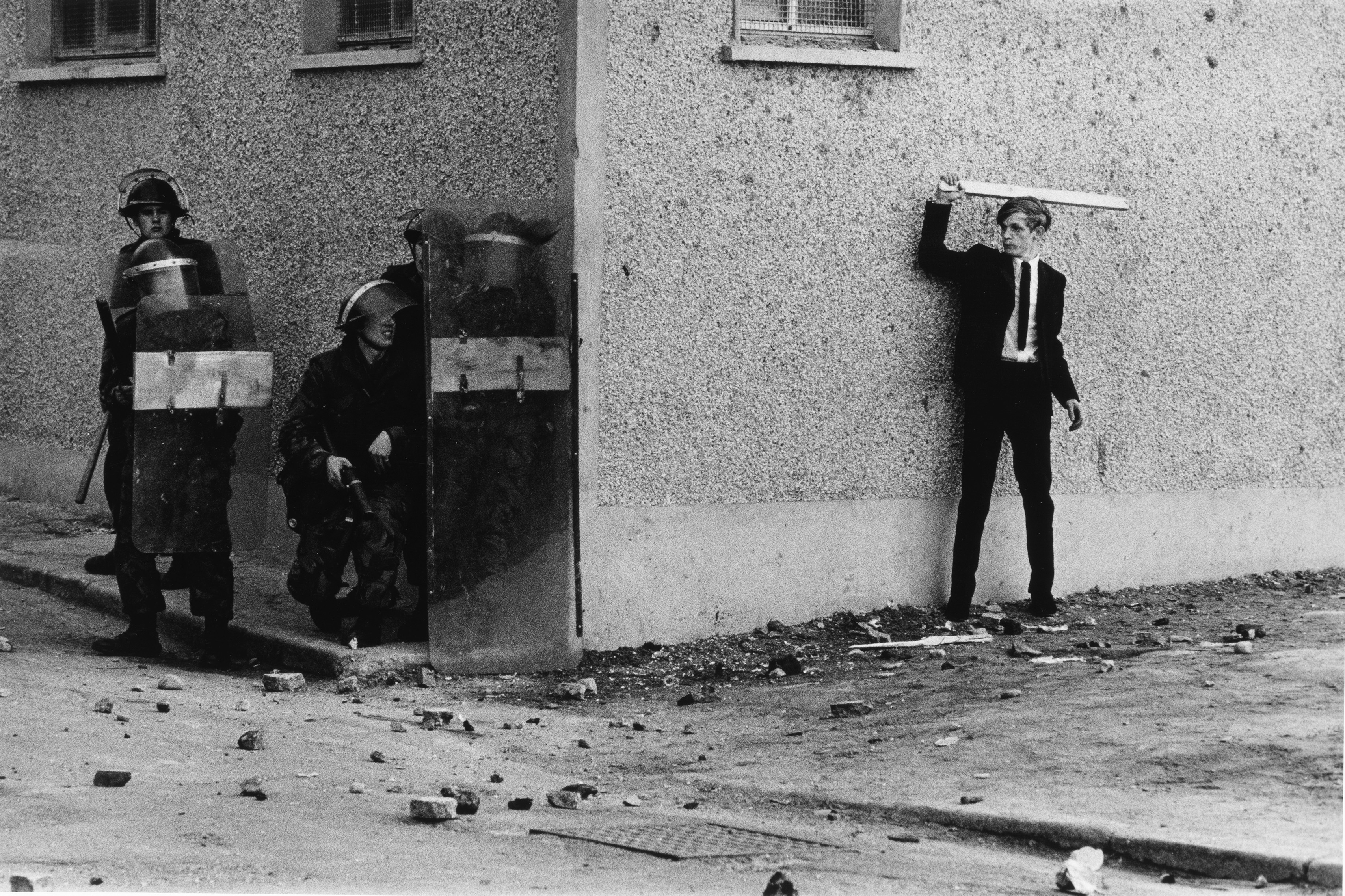 A young man in a suit and tie brandishes a stick at three police officers in riot gear and shields, in a street littered with debris.