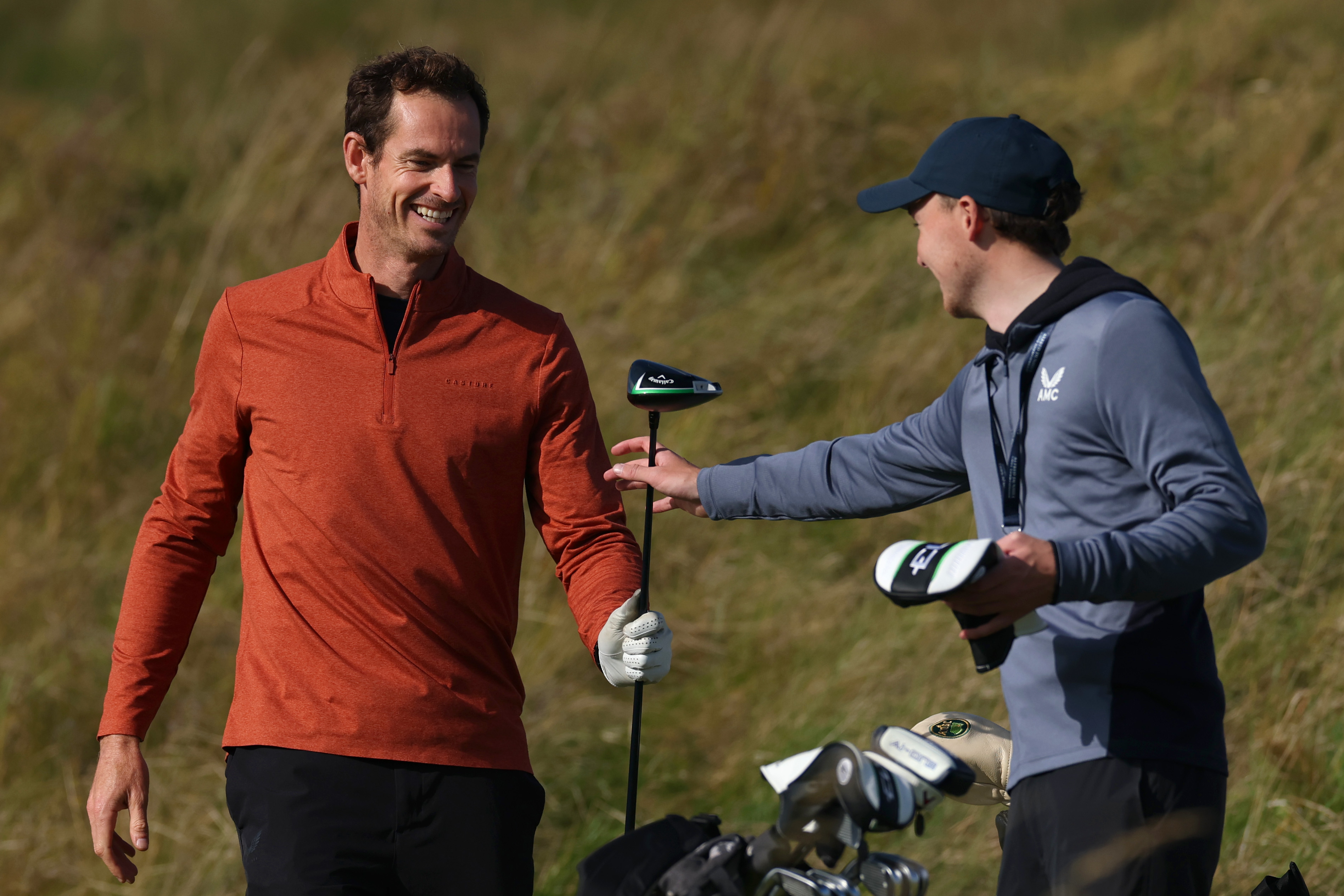 Andy Murray smiling and receiving a golf club from a caddie during practice.