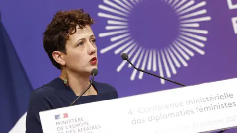 @judith.Litvine/MEAE A woman with short, dark, curly hair speaks at a conference. She is wearing a navy blue top with dangly earrings. A podium with two microphones is in front of her and a purple screen is behind her.  