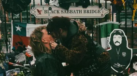 Sam Bok A couple kiss at the Black Sabbath Bridge. The man with long blonde hair and a black leather jacket is on his knees. He has just proposed. The woman has dark hair and a leopard print coat. We can see the Black Sabbath Bridge behind them. 
