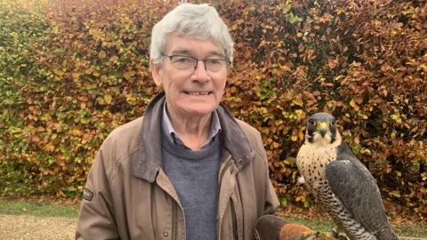 Contributed Mick Smith smiles at the camera as he holds his falcon Chanel on his arm while wearing a glove. Mick has grey hair and wears a brown coat, grey jumper and shirt. Chanel has some dark feathers on her wings and head, mixed with some lighter ones.
