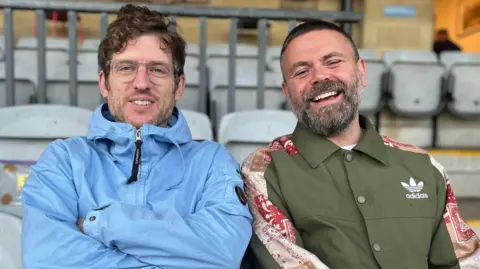 Geraint John Elis James are sitting in football stands, looking ointo the camera and laughing.  Geraint has short dark greying hair and a beard. He is wearing a green buttoned-up jacket with red detailing on the shoulder. Elis is wearing glasses with a  translucent rim and and light blue waterproof jacket with a hood. He has brown curly hair. 