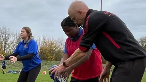 Tom Williams/BBC Three people on a rugby pitch. On the right is a man, sideways on, in a black kit standing beside a girl in a red bib over a blue short-sleeved shirt. They are both bending over and looking towards a rugby ball, held by the girl. The man also has a hand on the ball. On the left is a girl in a blue short-sleeved shirt holding out her hands ready to catch something. 