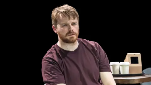 Johan Persson Jack Lowden sits in a chair on stage in The Fifth Step, looking concerned, and wearing a burgundy T-shirt.