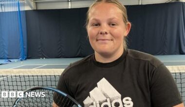 Ruby Bishop at a tennis court. She has blonde, swept-back hair and is wearing a black T-shirt with a large white Adidas logo. She is holding a tennis racket and looking direct at the camera.