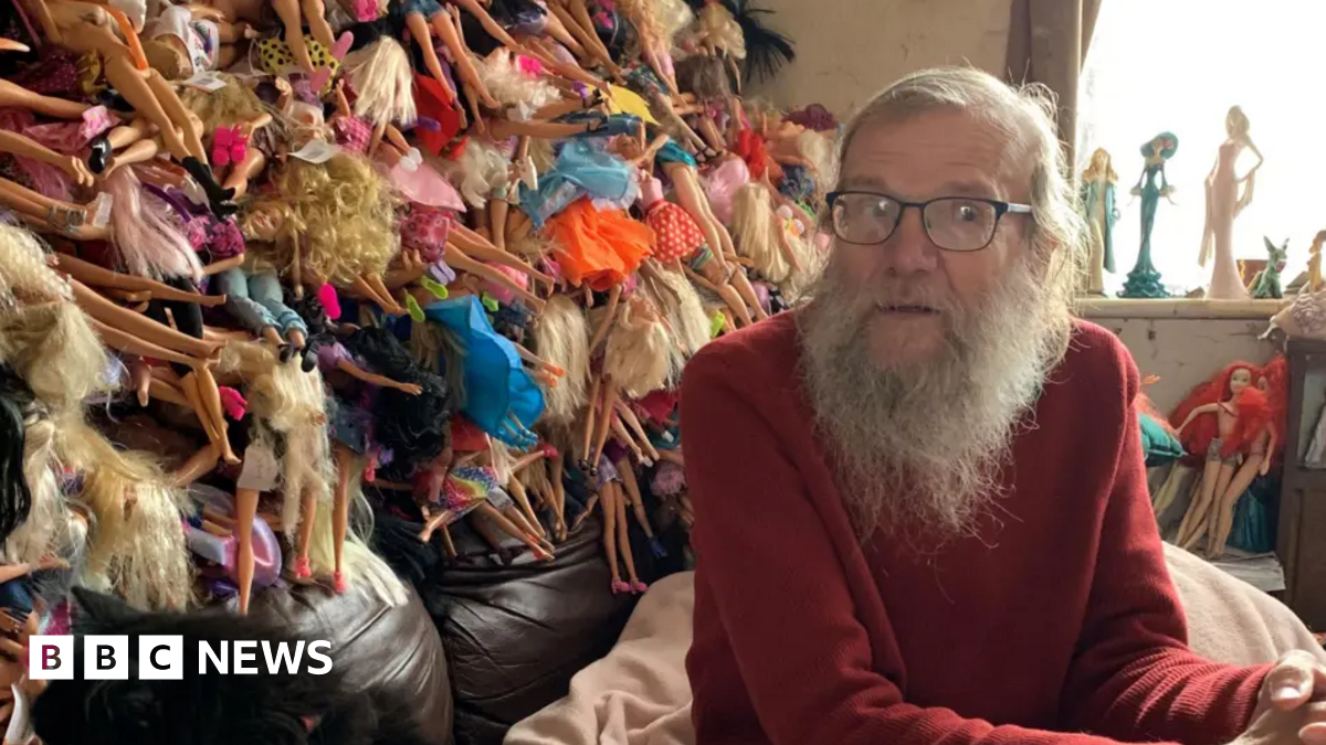David is sitting in front of a huge pile of Barbie dolls sticking out of several bin bags. He has long grey hair and a beard and glasses. He is wearing a red jumper.