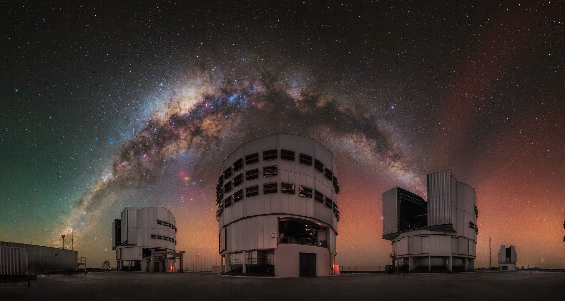 A glowing arch of the Milky Way galaxy can be seen in a red and orange and green night sky over three large towering buildings.