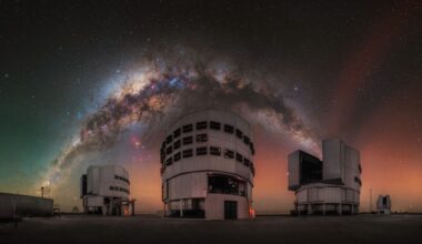 A glowing arch of the Milky Way galaxy can be seen in a red and orange and green night sky over three large towering buildings.
