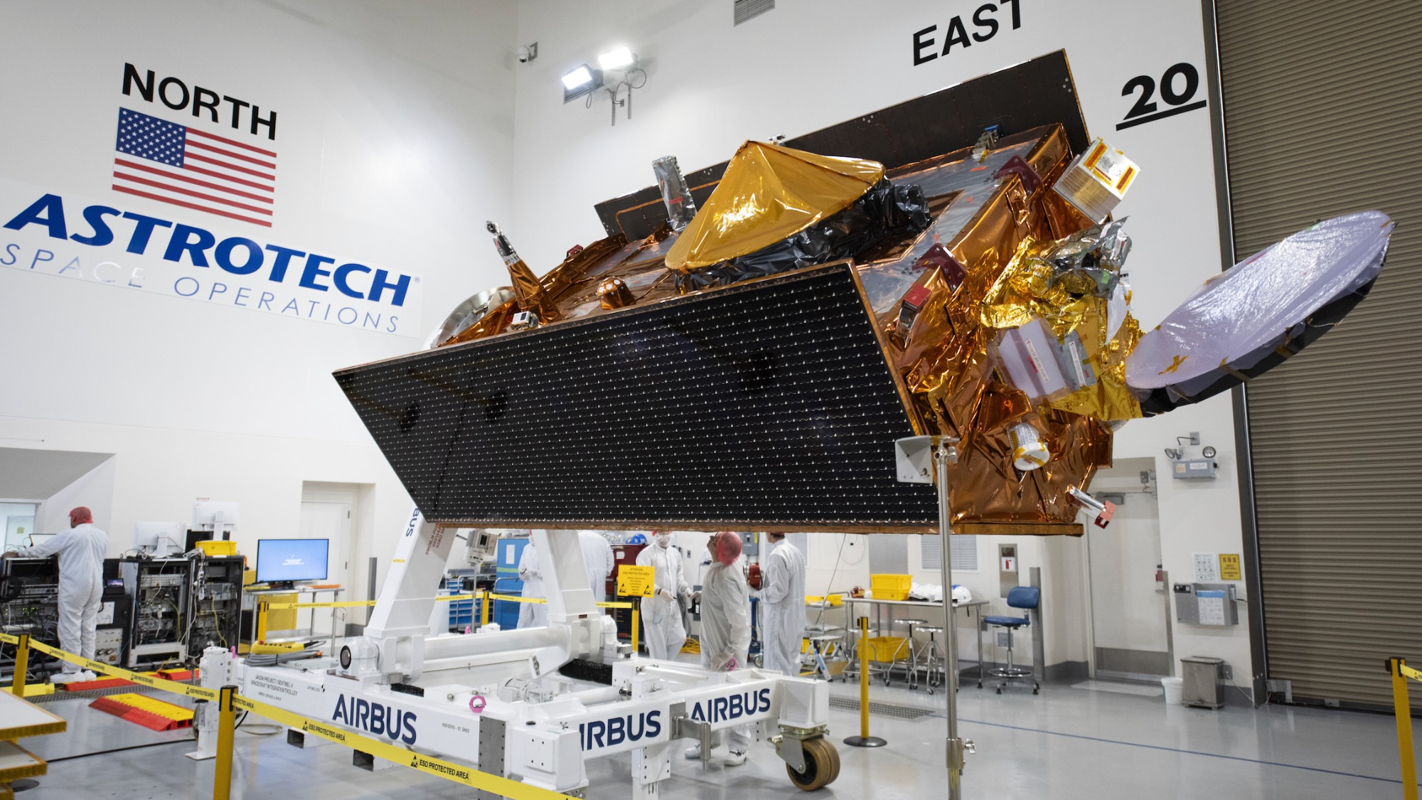 a large golden spacecraft sits in a white-walled clean room with technicians around it