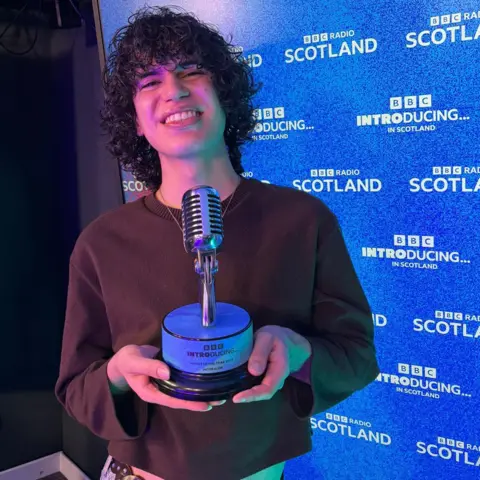 Jacob Alon has dark, curly hair and is smiling at the camera as they hold the award - shaped as a microphone. There a blue background saying BBC introducing in Scotland behind them