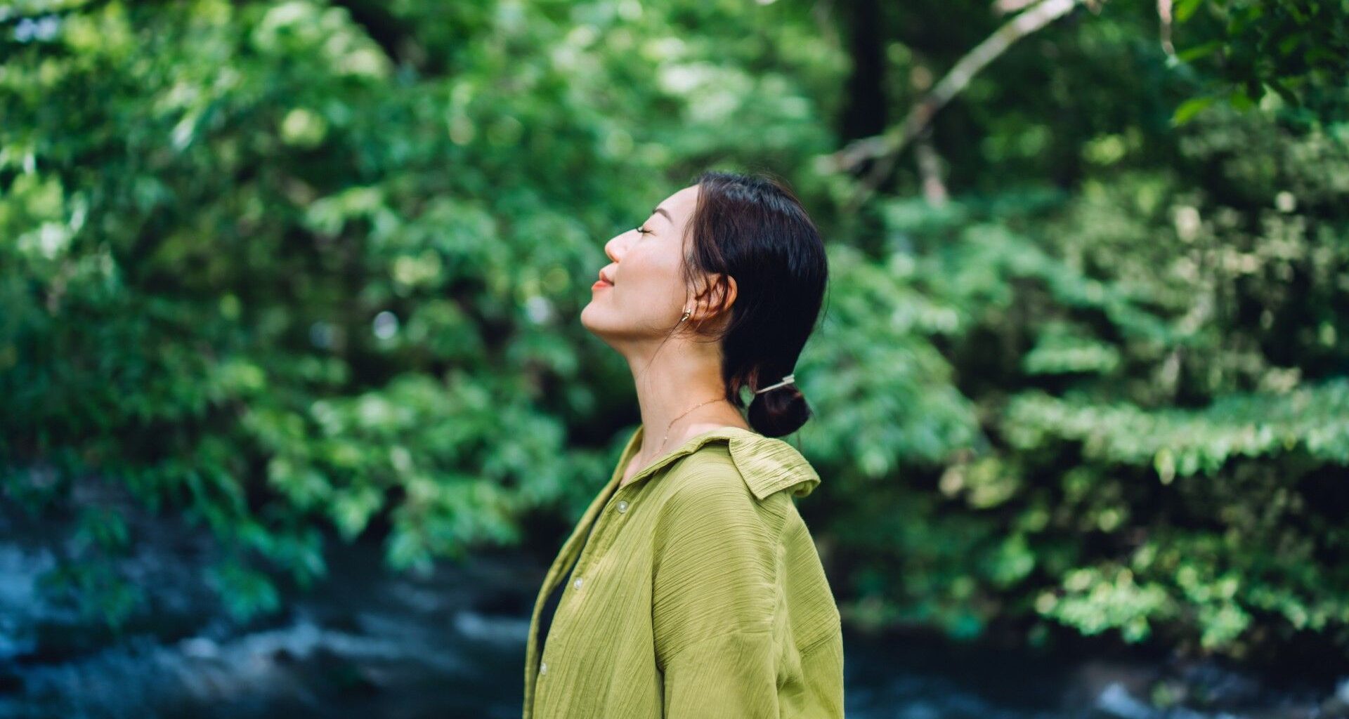 Woman in green blouse standing in nature with eyes closed smiling