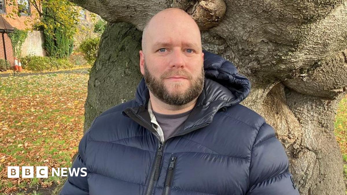 Pete White is an RAF veteran who left the armed forces in 2017. He is bald with a beard and is wearing a puffer jacket and standing in front of a tree, looking into the camera. There is a grassed area behind him.
