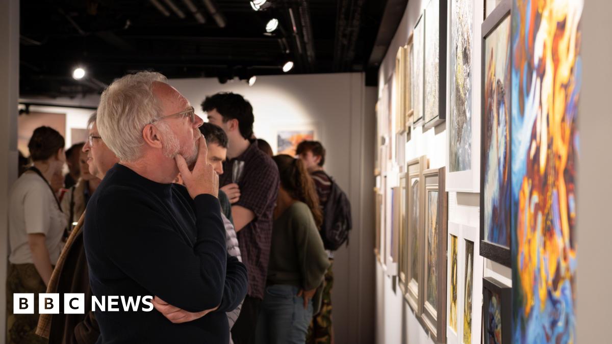 A man with grey hair, glasses and a dark jumper looks at the paintings on one of the gallery's walls. Behind him lots of people are milling around the gallery space.
