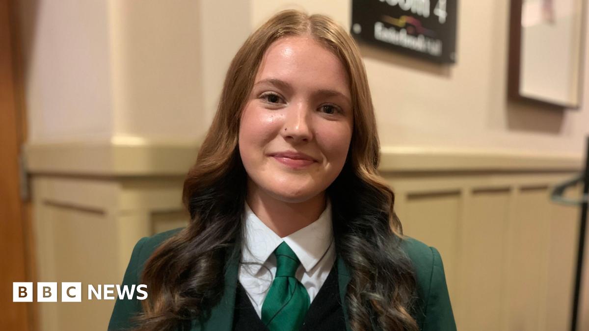 A young woman with long brown hair and a green school uniform on with green tie and a white blouse and green blazer