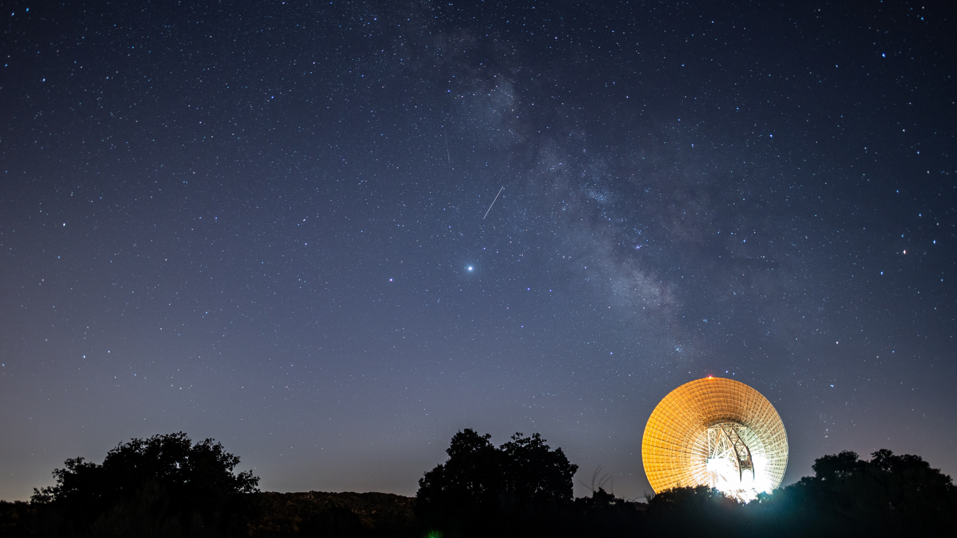 A night sky is pictured teeming with stars. The glowing band of the Milky Way is glowing diagonally across the image. It meets the illuminated disk of a large radio telescope near the horizon, which is silhouetted by trees.