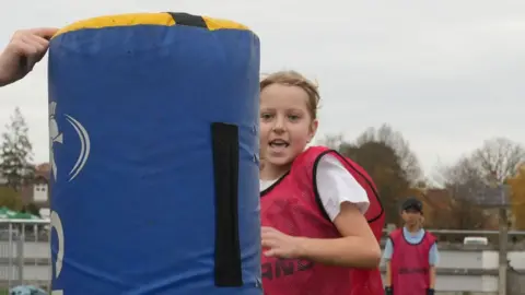 John Fairhall/BBC A primary school-aged girl in a pink tabard over a white T-Shirt running towards and about to make contact with a blue tackle bag, an upright foam pad about 1.5 metres (4.9ft) tall.  Behind her is another, similarly-dressed pupil. They are on the grounds of a rugby pitch.