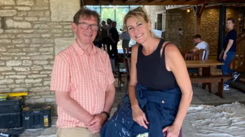 Martin Taylor A man with short brown hair and wearing an orange shirt is standing with his hands clasped in front of him next to a woman with tied-back blonde hair and wearing a black vest. Both are smiling at the camera. There are people standing and sitting inside a barn-type building behind them. 