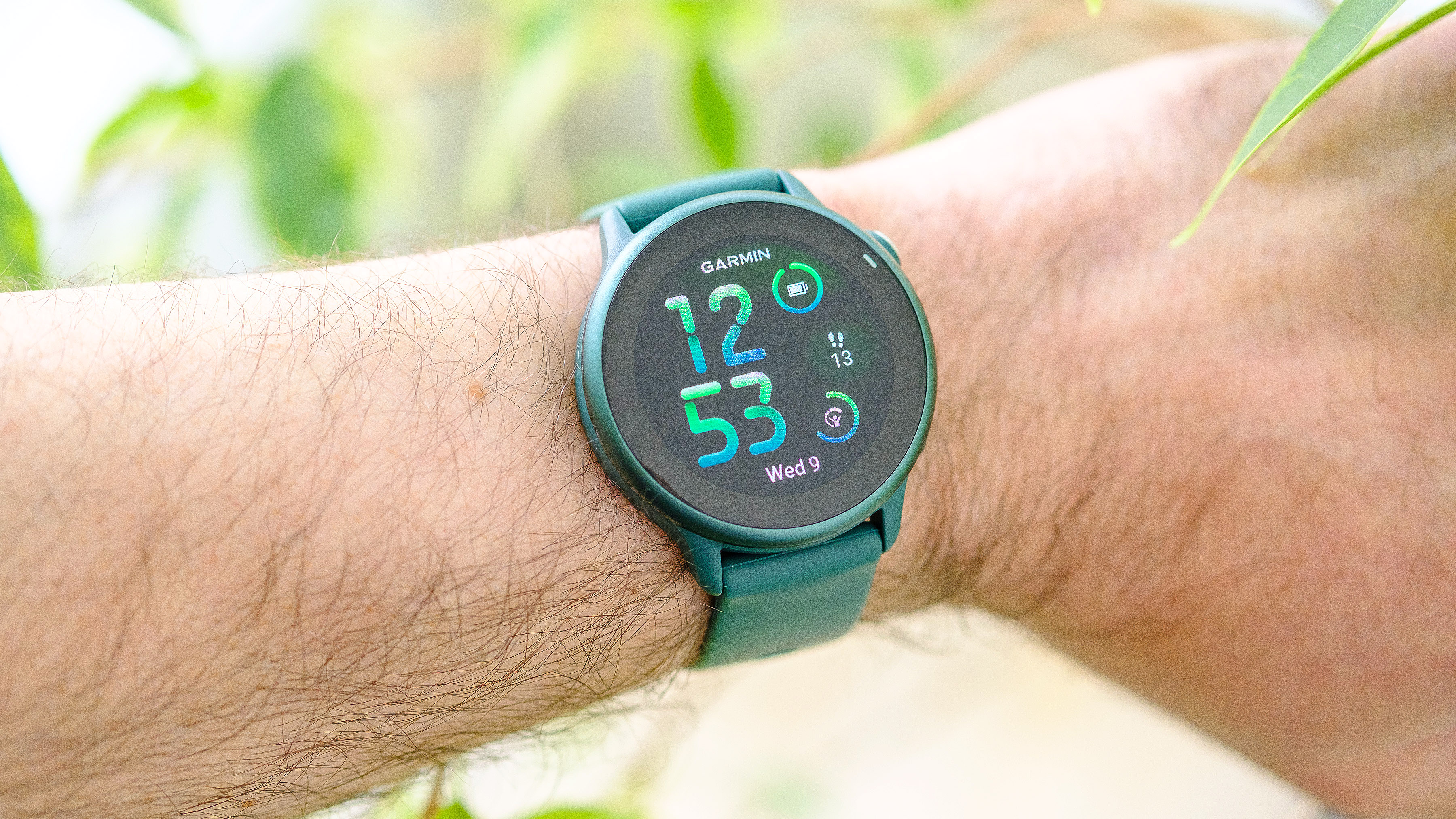 Close-up of the Garmin Vivoactive 6 smartwatch in green on a user's wrist with foliage shown out of focus around it