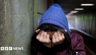 A woman wearing a blue hoodie and a purple jacket with her face buried in her hands, standing in an alleyway.