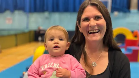On the left is a one-year-old baby with blonde hair. She has her mouth open and is lookingt at the camera. She is wearing a pink jumper with the word friends on it. On the right is Keri Rang, who has brown shoulder length hair. She is wearing a black jumper and blouse with a silver necklace with a circular charm on the end.