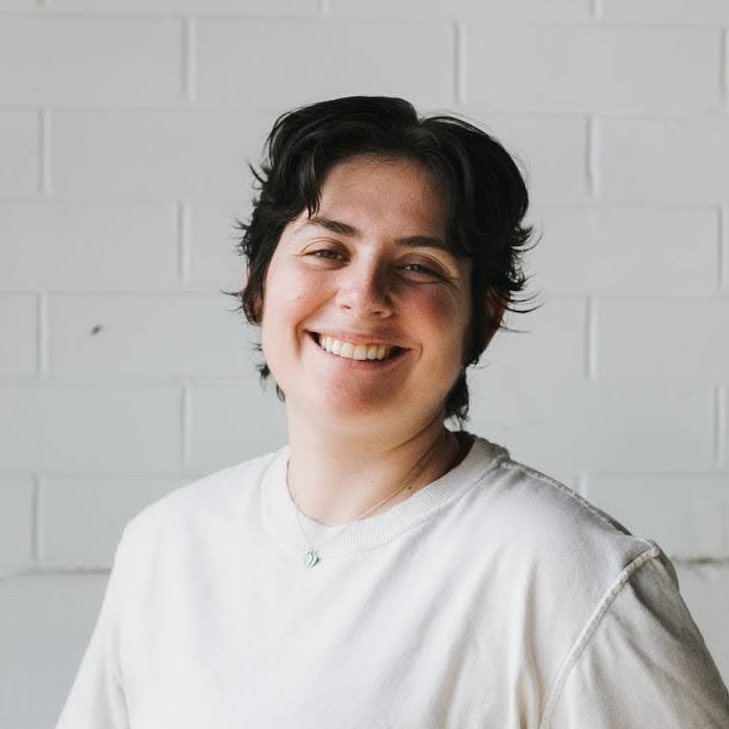 Head and shoulders of smiling person with short black hair wearing white T-shirt, in front of white brick wall