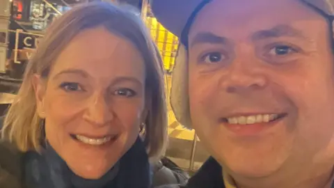 Geraint John Deb and Geraint smiling at the camera. She has a short blonde bob and he is wearing a cap with ears.