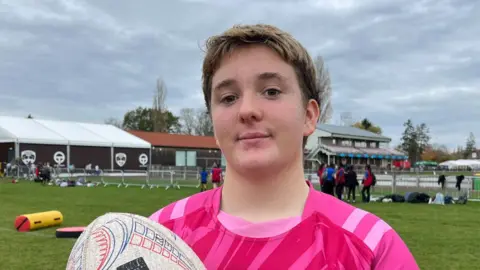 Tom Jackson/BBC A mid-teenage girl with short hair and wearing a pink rugby shirt and holding a white rugby ball. She is standing on a pitch and behind her can be seen club buildings. 