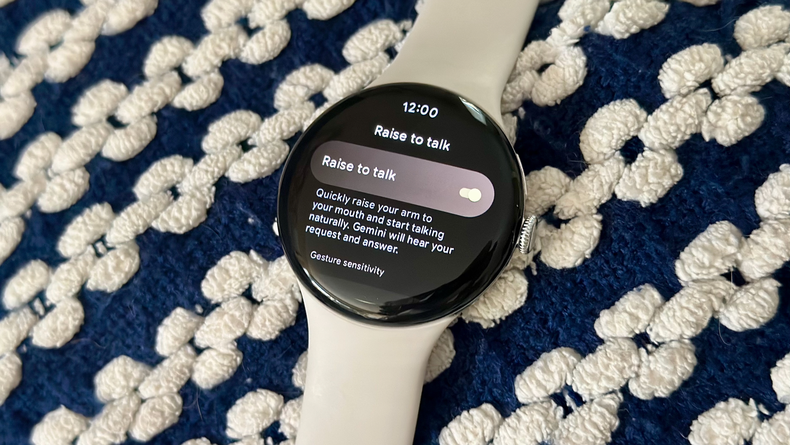 A photo of the Google Pixel Watch 4 on a blanket showing the "Raise to Talk" settings menu, with the feature toggled on.