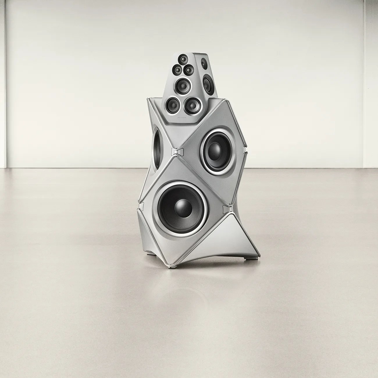 Silver multi-driver speaker system with a geometric design standing on a smooth floor against a plain wall.