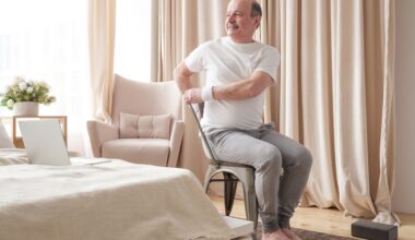 man in a white tshirt and grey joggers sitting in a chair and twisting to one side. he's in a bedroom setting with a laptop on the bed and a yoga block on the floor.