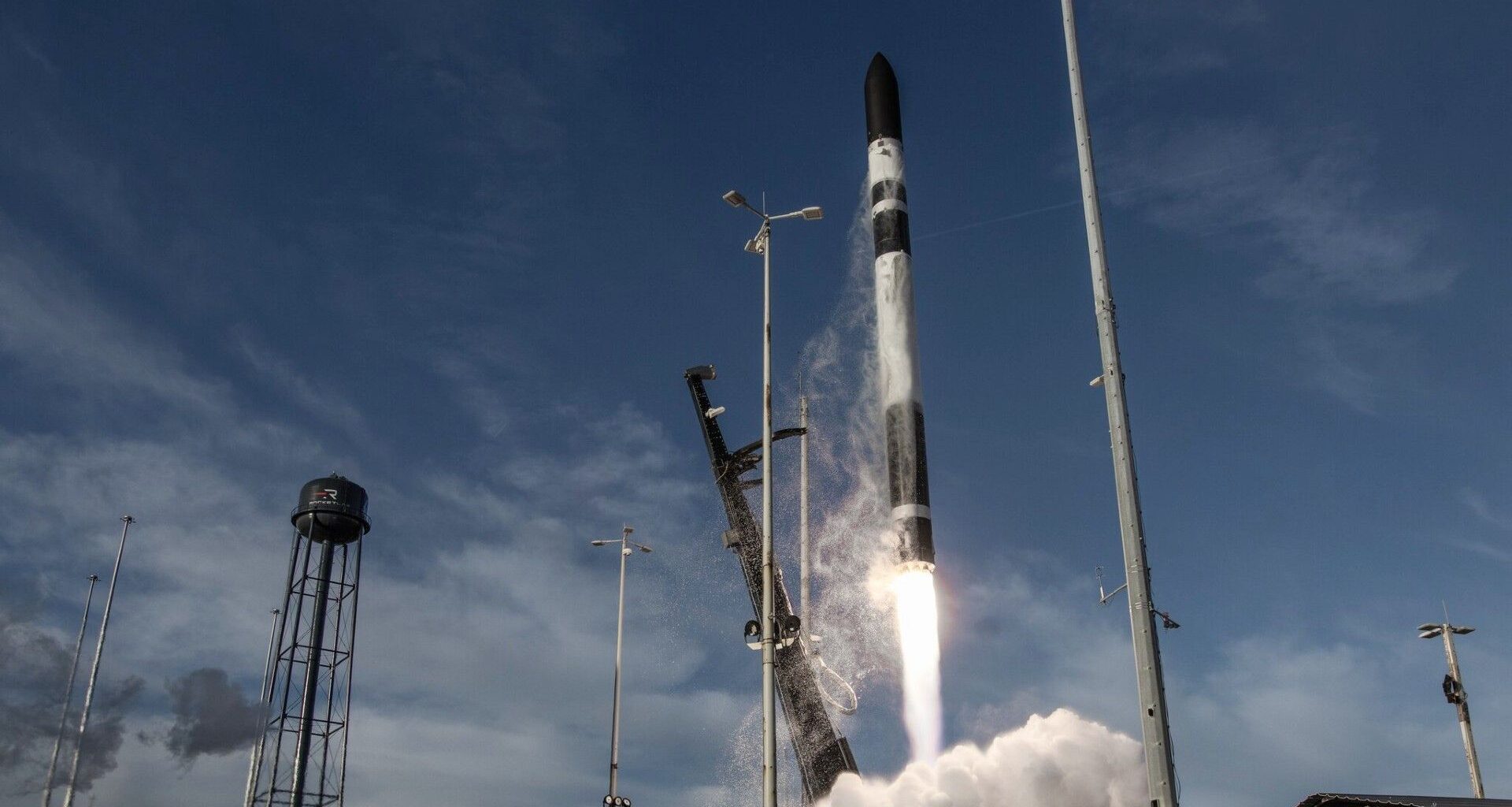 a black and white rocket lifts off above a cone of fire and smoke next two a black water tower