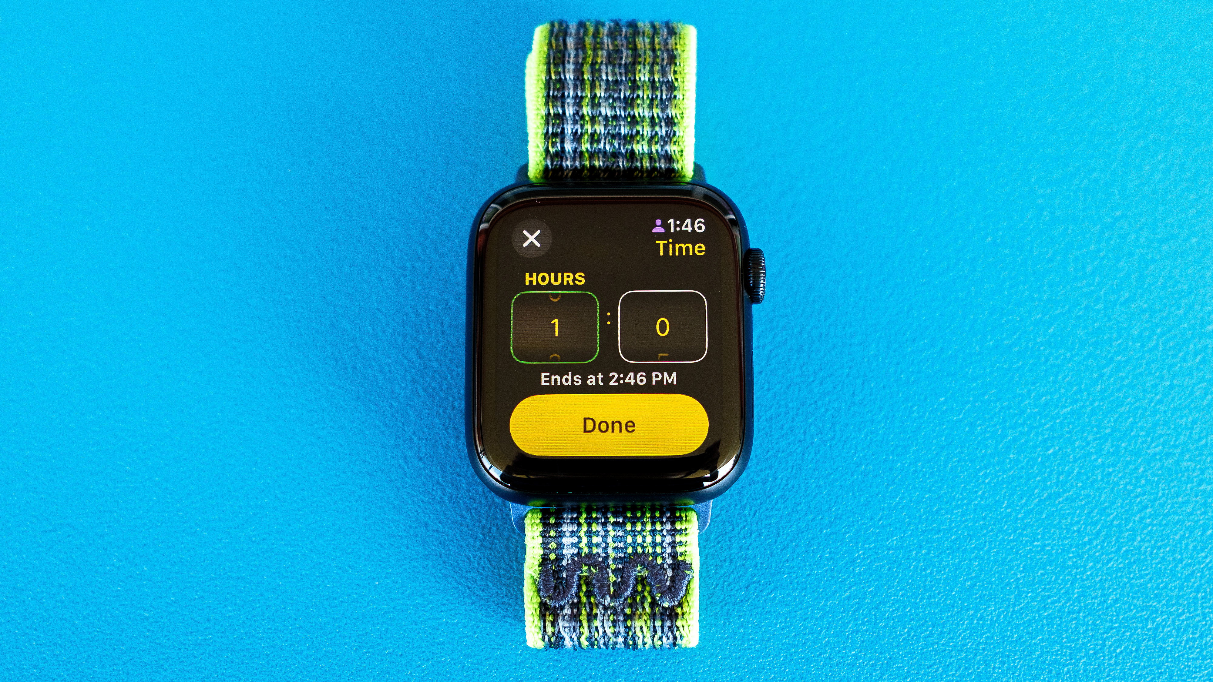Detail shot of an Apple Watch SE on a bright blue background showing the steps to set up and send a Check In notification in watchOS 11
