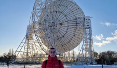A young man in a red jacket standing in the snow with a radio dish behind him.