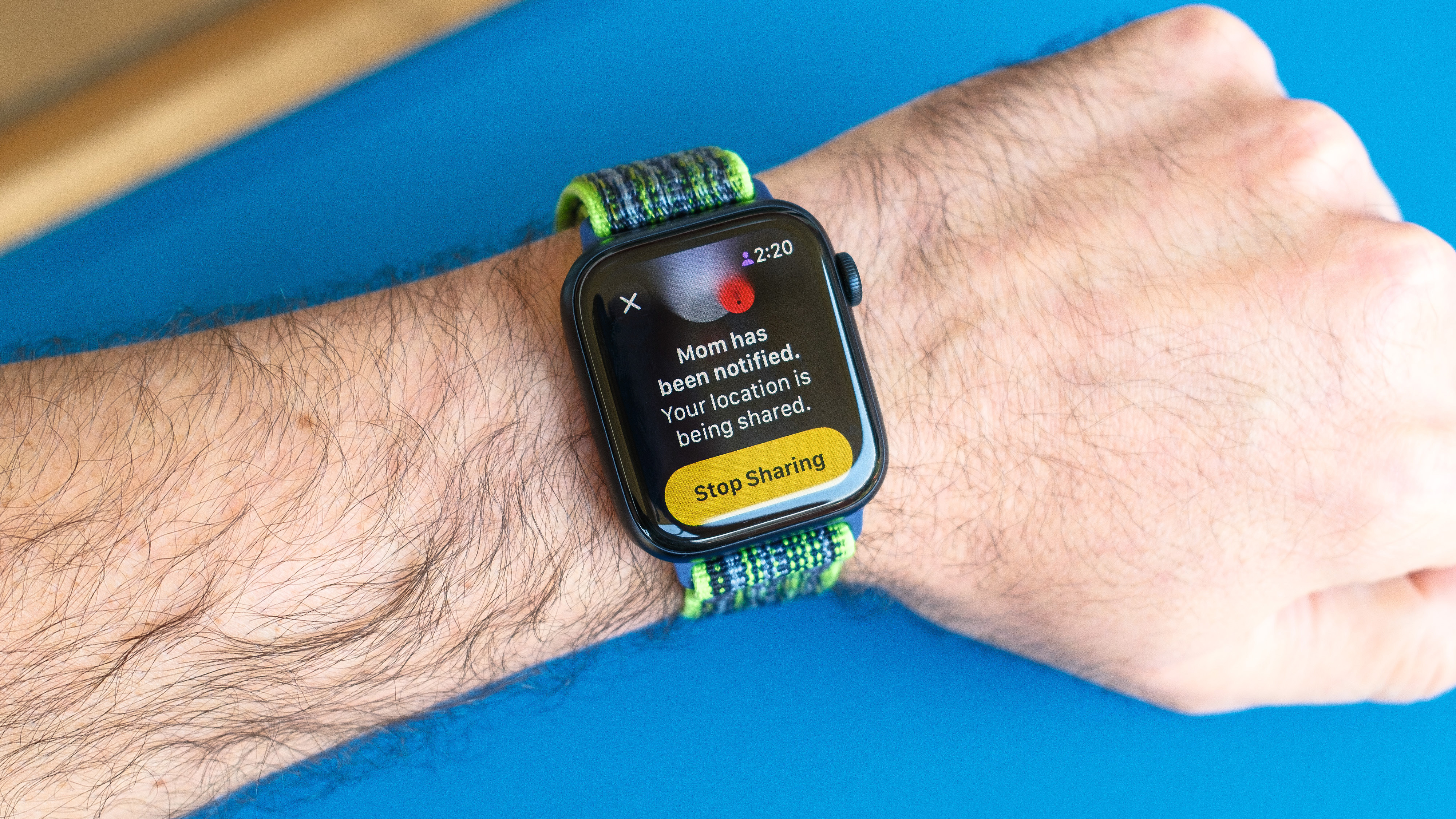 Detail shot of an Apple Watch SE on a bright blue background showing the steps to set up and send a Check In notification in watchOS 26