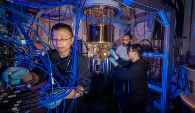 Three researchers work in a small lab space full of blue wires and a golden dilution fridge.