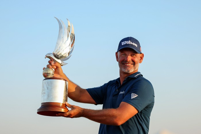 Paul Waring, in blue Nike shirt and cap, smiles as he holds the Abu Dhabi Golf Championship 2024 trophy.