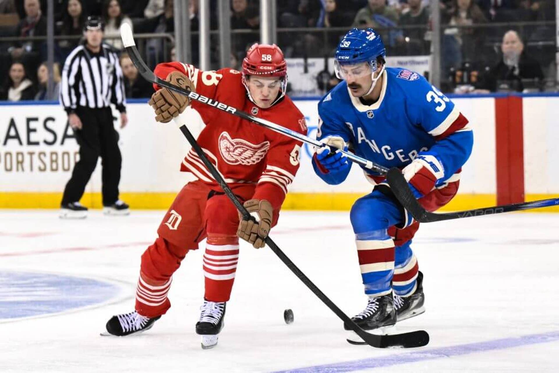 A Red Wings player in a red jersey battles for the puck against a Rangers player in a blue jersey during an NHL game.