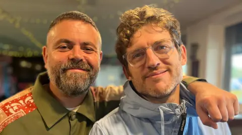 BBC Geraint John has his arm around Elis James and both are smiling and looking at  the camera. Geraint has short dark greying hair and a beard. He is wearing a green buttoned-up jacket with red detailing on the shoulder. Elis is wearing glasses with a  translucent rim and and light blue waterproof jacket with a hood. He has brown curly hair. 