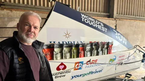 BBC Ollie Phillips smiling at the camera is wearing a burgundy jumper and black gilet with a rowing boat in the background.