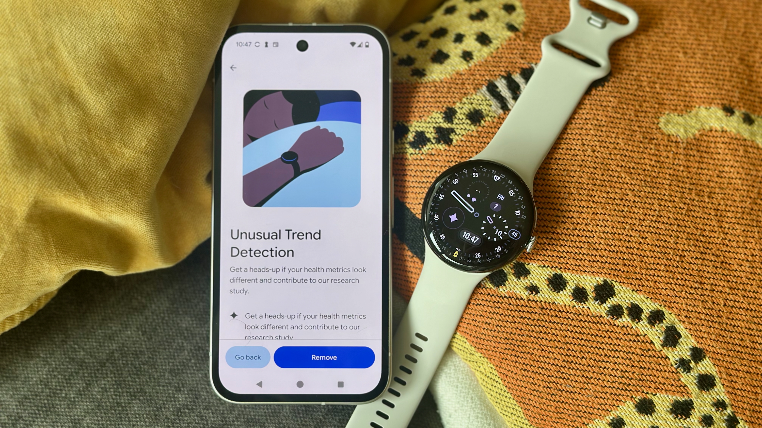 A photo of an Android phone showing an "Unusual Trend Detection" sign-up page in the Fitbit app for Fitbit Labs, sitting next to a Google Pixel Watch 4 atop colorful pillows.