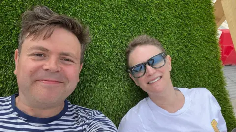 Geraint John Geraint and Deb lying on grass. Geraint is wearing a navy and white stripy t-shirt and has short brown hair. Deb  is wearing a white t-shirt and sunglasses and has her fair fair tied up. 