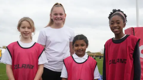 John Fairhall/BBC Four primary school girls in a row. The three in front are wearing deep pink tabards over white T-shirts and the one behind is wearing a white T-shirt. They are all smiling broadly and the rugby pitch can be glimpsed behind. 